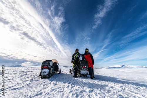 Two snowmobiles (back side) and two friends pose around of one snowmobile. They dressed for driving in cold mountains, waiting to start the holiday adventure. Hemavan, Tarnaby in Lappland, Sweden