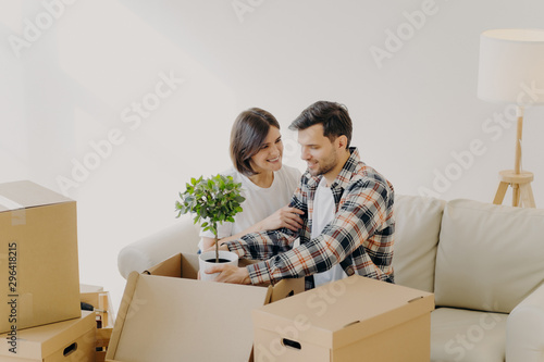 Happy couple in love settle in new home, unpack boxes with household objects, man holds potted plant, woman embraces and expresses love to husband, start new family life in modern apartment. © VK Studio
