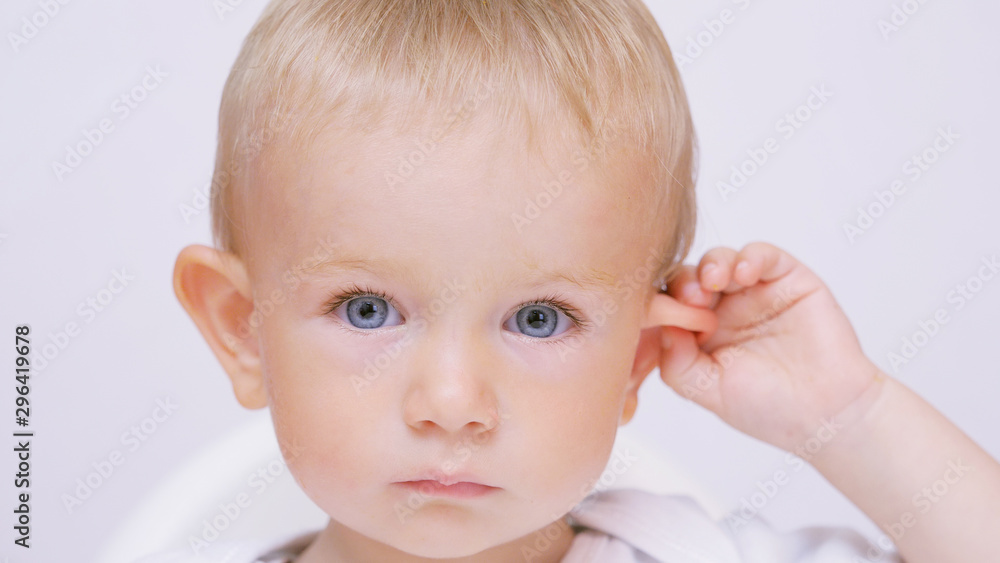 Blue eyes baby close up portrait, child play with ear, smiling face, purity