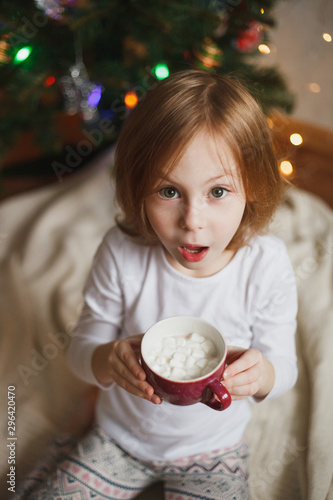 Funny surprised girl holding a cup of cocoa with marshmallows near a Christmas tree with lights.