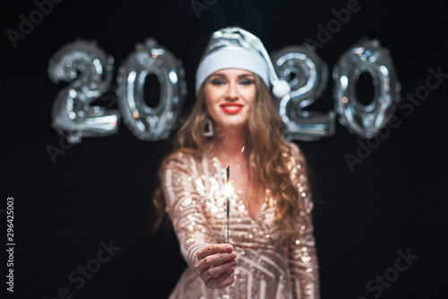Happy young woman in Santa hat with sparklers in hand against metallic 2020 balloons.
