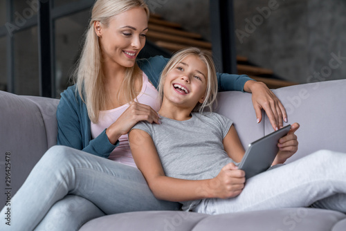 Happy mother and daughter having fun and using digital tablet sitting on sofa in living room