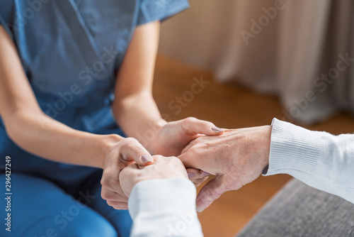 Assisting senior people. Close up of female caregiver holding elderly woman's hands indoors photo