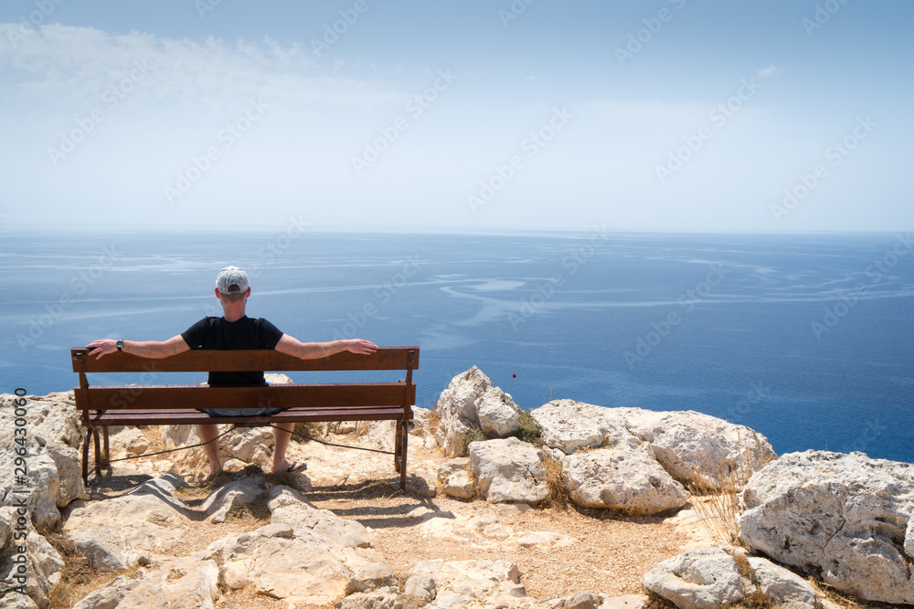 Guy sitting on wooden bench and looking at the blue ocean in Ayia Napa, Cape Greco National Forest Park, Cyprus Guy sitting on wooden bench and looking at the blue ocean in Ayia Napa, Cape Greco National Forest Park, Cyprus