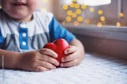 Smiling little child boy holding red toy heart in hands near window in daylight with cozy bokeh on background. The concept of love for children and cardiological care.