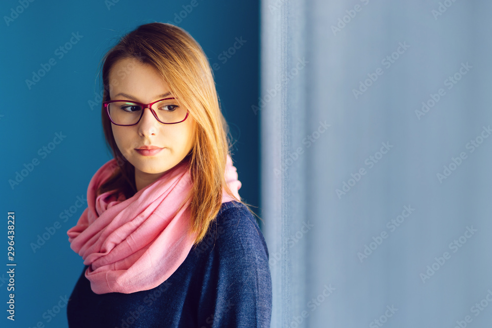 Portrait of young woman brunette wearing blue sweater and pink scarf in front of a blue wall by the window at home or work wearing glasses looking away Portrait of young woman brunette wearing blue sweater and pink scarf in front of a blue wall by the window at home or work wearing glasses looking away