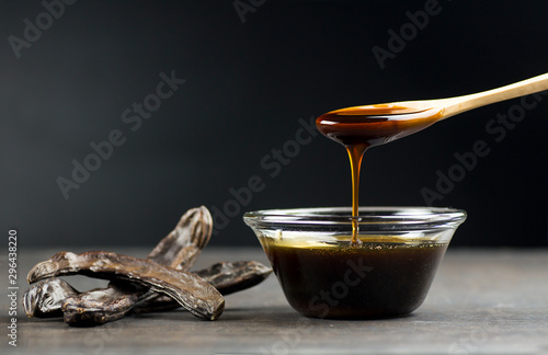 carob molasses in glass bowl and in wooden spoon and carob pods on rustic background, locust bean healthy food, Ceratonia siliqua ( harnup )