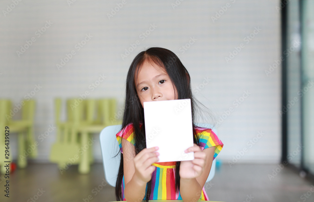 little Asian child girl holding blank white paper card in her hand. Kid showing empty paper note copy space in children room.