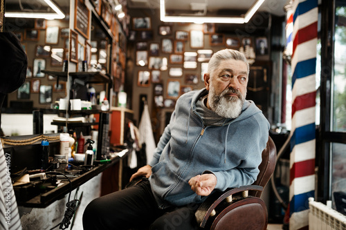A white haired bearded hipster man sits in a barbershop and waits for his turn to get a haircut