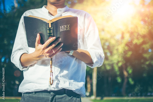 The young Asian man is reading the Holy Bible and the cross necklace, a symbol of redemption for the Man of Jesus, which he put on the center of book, amidst the atmosphere of the park with copy space