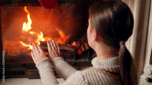 4k footage of young woman in sweater warming and rubbing her hands at burning fireplace in house photo