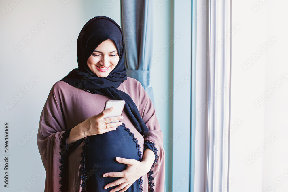 Ταπετσαρία Young pregnant arab woman in hijab using her mobile phone to send message