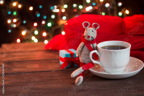 Christmas coffee on a dark wooden table. Blurred garland lights in the background. Freshly brewed coffee in a white cup. Good New Year spirit. Symbol of the year 2020 toy rat.