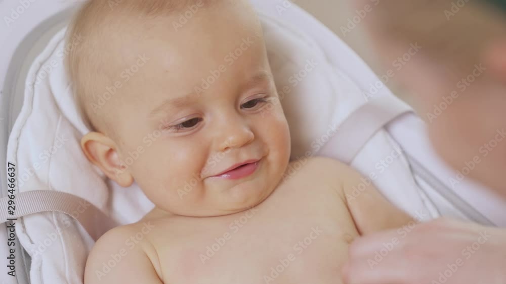 A close-up of a sophisticated mother doing gymnastic to her adorable babies hands to reduce muscle tone while her son is lying in baby seat. Child body-care. Selective focus shot A close-up of a sophisticated mother doing gymnastic to her adorable babies hands to reduce muscle tone while her son is lying in baby seat. Child body-care. Selective focus shot