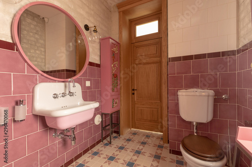 Interior of clean light restroom with retro sink beside big round mirror and decorated cabinet on tall wall with white and pink tile next to toilet bowl with brown lid in apartment