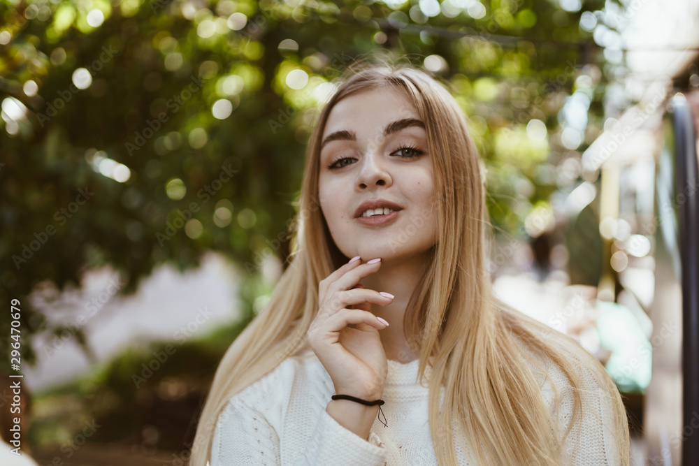 Gorgeous young girl with blond long straight hair in the white knitted sweater stands against the backdrop of a city park and poses, smiles, enjoys the nature in the sunshine behind her. Gorgeous young girl with blond long straight hair in the white knitted sweater stands against the backdrop of a city park and poses, smiles, enjoys the nature in the sunshine behind her.