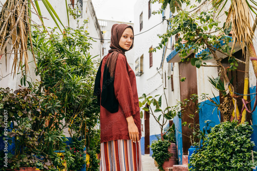 Charming girl with a terracotta scarf on her head and a brown blouse in striped trousers stands in the courtyard of the eastern courtyard of white-blue color with greenery Charming girl with a terracotta scarf on her head and a brown blouse in striped trousers stands in the courtyard of the eastern courtyard of white-blue color with greenery
