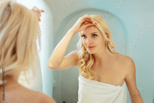 blond woman in a white bathroom looking at her reflection in the mirror, wearing a towel