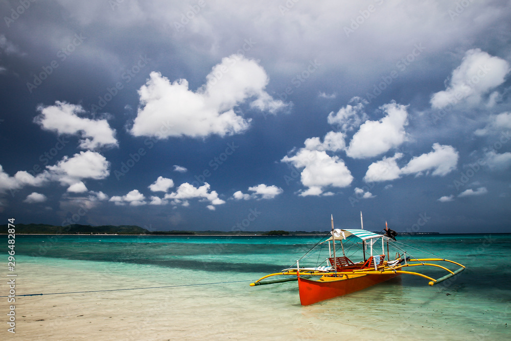 bateau de pêche coloré orange et jaune sur mer turquoise et sable blanc accosté sur une île aux Philippines sur fond de ciel d'orage gris et nuages blancs
