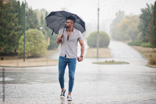 Fashionable tall arab beard man wear on shirt, jeans and sunglasses with umbrella posed at rain on park square.