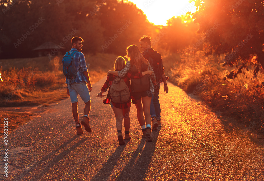 Group of four friends having fun hiking through countryside together on road during sunset. - obrazy, fototapety, plakaty 
