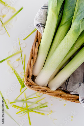 Fresh leeks in a wooden brown basket on top of a  white tablecloth and fresh leek cuttings on a white background