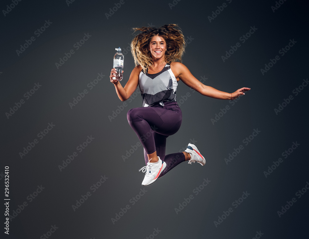 Exited cheerful woman is jumping at studio while holding the bottle of water over grey background.