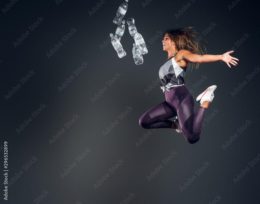 Happy smiling woman trow away bottles of water while jumping at grey studio background.