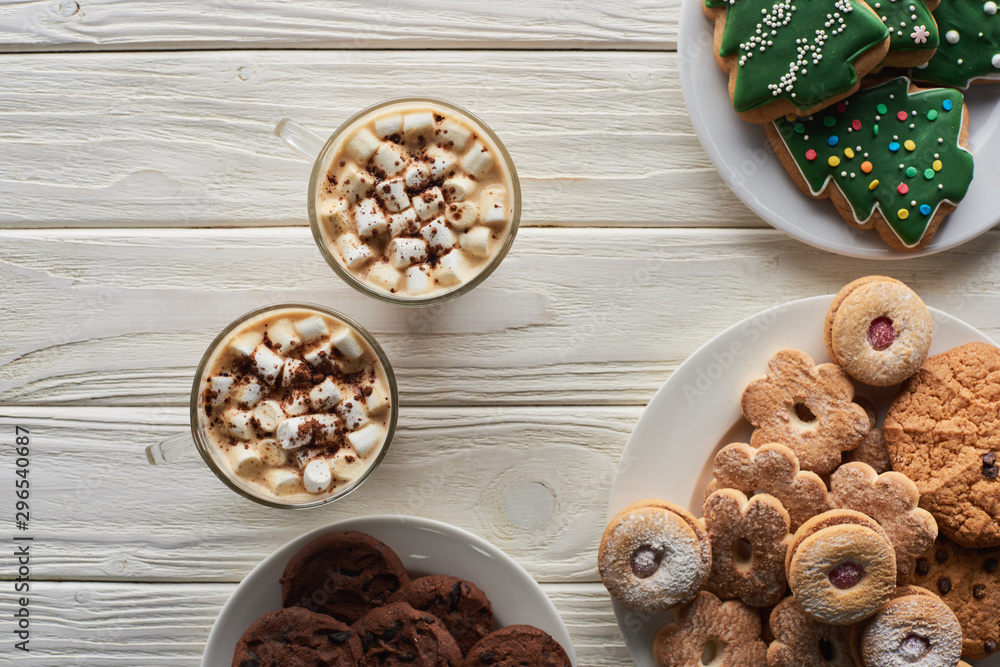 top view of cacao with marshmallow and cacao powder in mugs near Christmas cookies on white wooden table top view of cacao with marshmallow and cacao powder in mugs near Christmas cookies on white wooden table