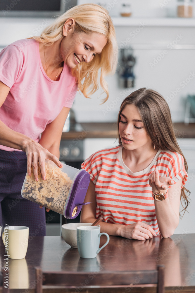 smiling mature woman adding flakes in bowl while young daughter sitting at kitchen table smiling mature woman adding flakes in bowl while young daughter sitting at kitchen table