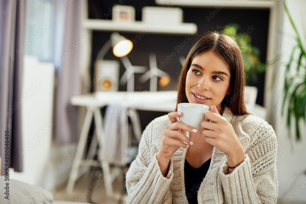 Caucasian woman dressed in beige sweater sitting on bed and holding fresh morning coffee. Caucasian woman dressed in beige sweater sitting on bed and holding fresh morning coffee.
