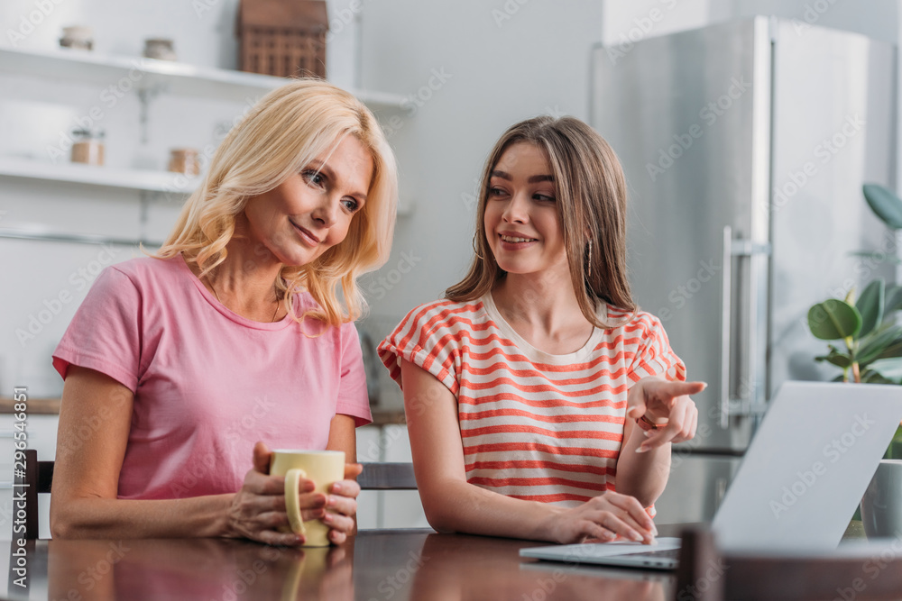 cheerful girl pointing with finger at laptop while sitting at kitchen table near smiling mother cheerful girl pointing with finger at laptop while sitting at kitchen table near smiling mother