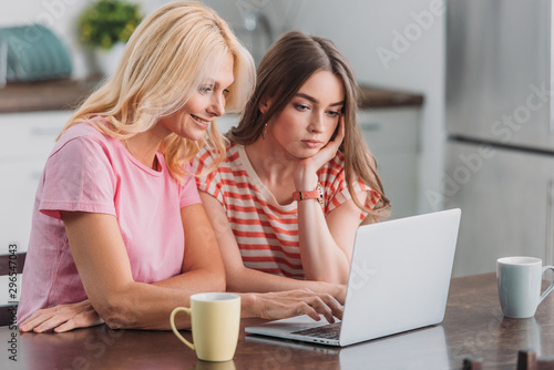 attentive girl sitting at kitchen table near smiling mother using laptop