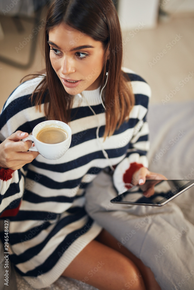 Beautiful caucasian brunette dressed in sweater sitting on bed in bedroom, using tablet and drinking coffee in morning. Beautiful caucasian brunette dressed in sweater sitting on bed in bedroom, using tablet and drinking coffee in morning.