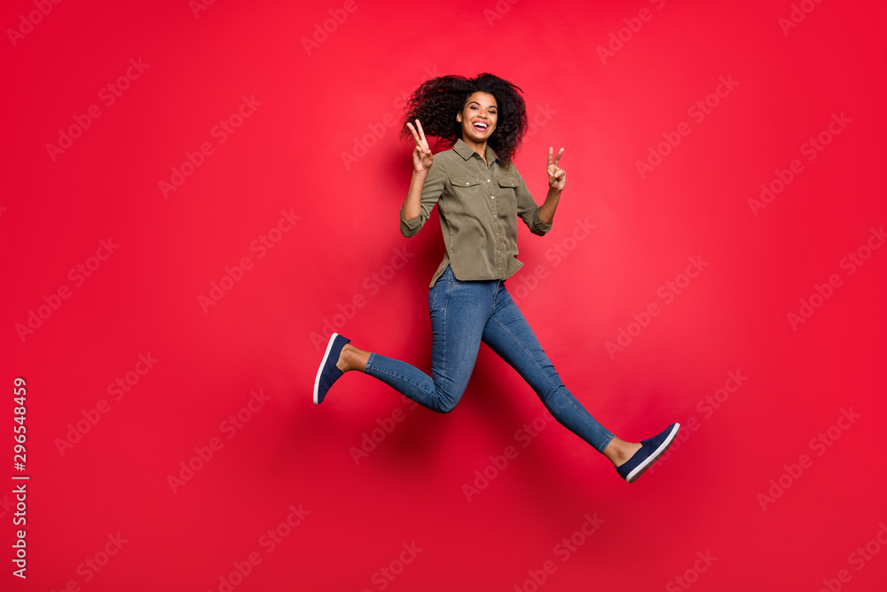 Full length body size photo of cheerful positive curly wavy brunette funky girlfriend running jumping showing v-sign wearing jeans denim shirt smiling toothily isolated vivid color background