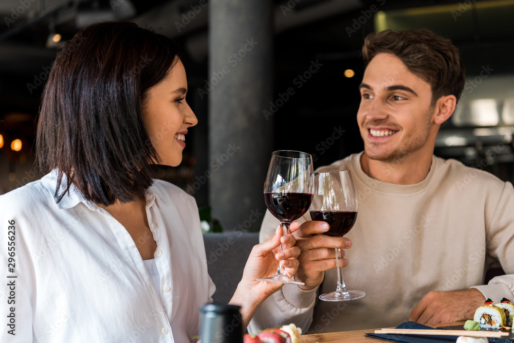 happy man and cheerful woman clinking glasses with red wine near sushi