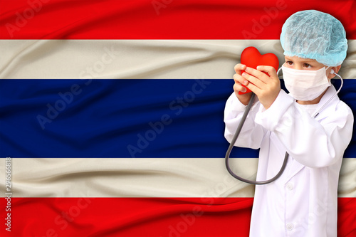 child, boy, in a white doctor’s coat, hat and mask attached a stethoscope to a red heart model, Thailand flag background, close-up, focus on the face, medical concept, cardiology, copy space