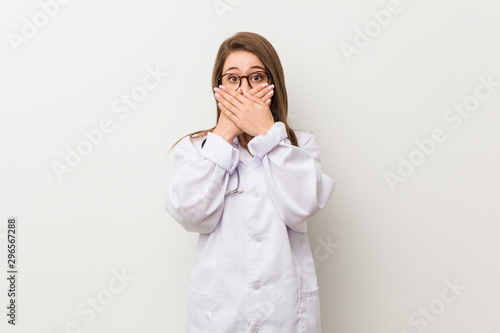 Young doctor woman against a white wall shocked covering mouth with hands.