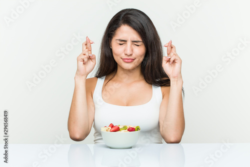 Young hispanic woman eating a fruit bowl crossing fingers for having luck photo