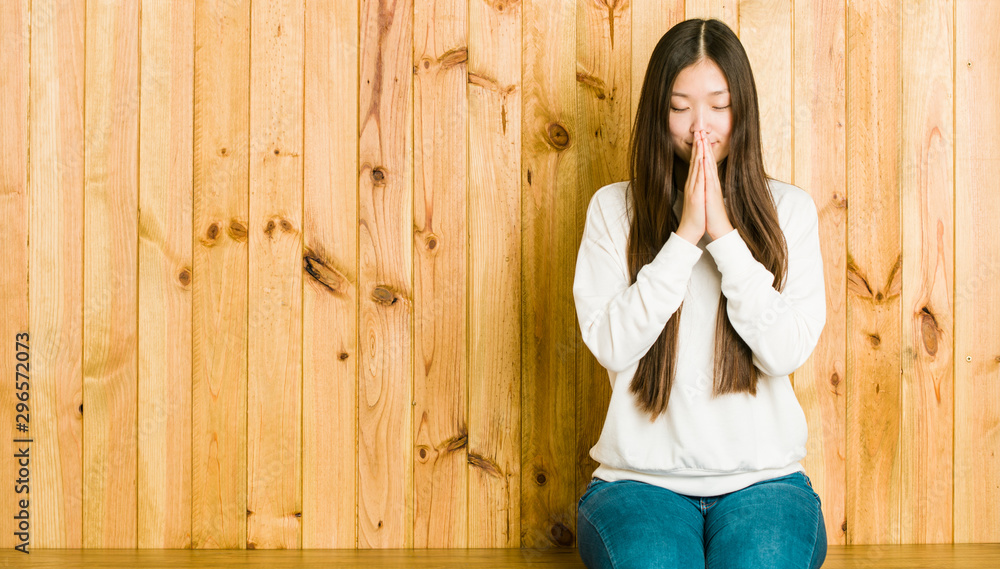Fototapeta premium Young chinese woman sitting on a wooden place holding hands in pray near mouth, feels confident.