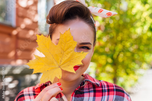 Beautiful woman with make up and hair in pin up style standing on the street, holding yellow maple leaf close to her face. photo