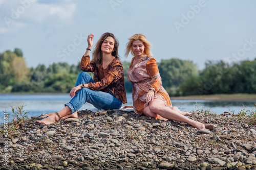 Two pretty sisters sitting on the small stone river island on a cloudy windy summer day, discuss girls secrets, laugh.