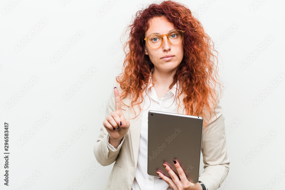 Young caucasian business redhead woman holding a tablet showing number one with finger. Young caucasian business redhead woman holding a tablet showing number one with finger.