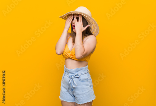 Young caucasian woman wearing a straw hat, summer look blink through fingers frightened and nervous.