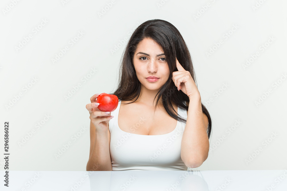 Young hispanic woman holding a tomato pointing his temple with finger, thinking, focused on a task. Young hispanic woman holding a tomato pointing his temple with finger, thinking, focused on a task.