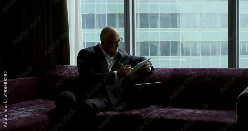 A bald businessman in a business suit and glasses, he sits in the room at the panoramic window on a purple sofa, on his knees he has a laptop, he holds a notebook with a pen, writes. A bald businessman in a business suit and glasses, he sits in the room at the panoramic window on a purple sofa, on his knees he has a laptop, he holds a notebook with a pen, writes.