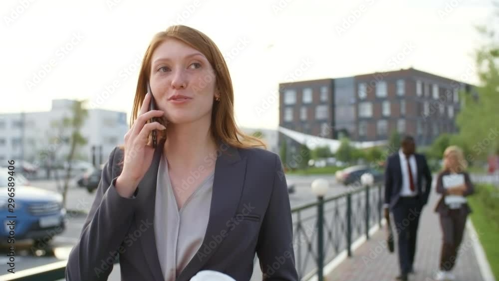 Front view medium shot of young ginger haired businesswoman walking on street, holding cup of takeaway coffee and having telephone conversation Front view medium shot of young ginger haired businesswoman walking on street, holding cup of takeaway coffee and having telephone conversation