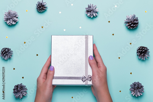 Woman manicured hands holding silver giftbox on blue background with pine cones and confetti, copy space, top view, flat lay. Background for Christmas or New Year. Giving presents concept photo