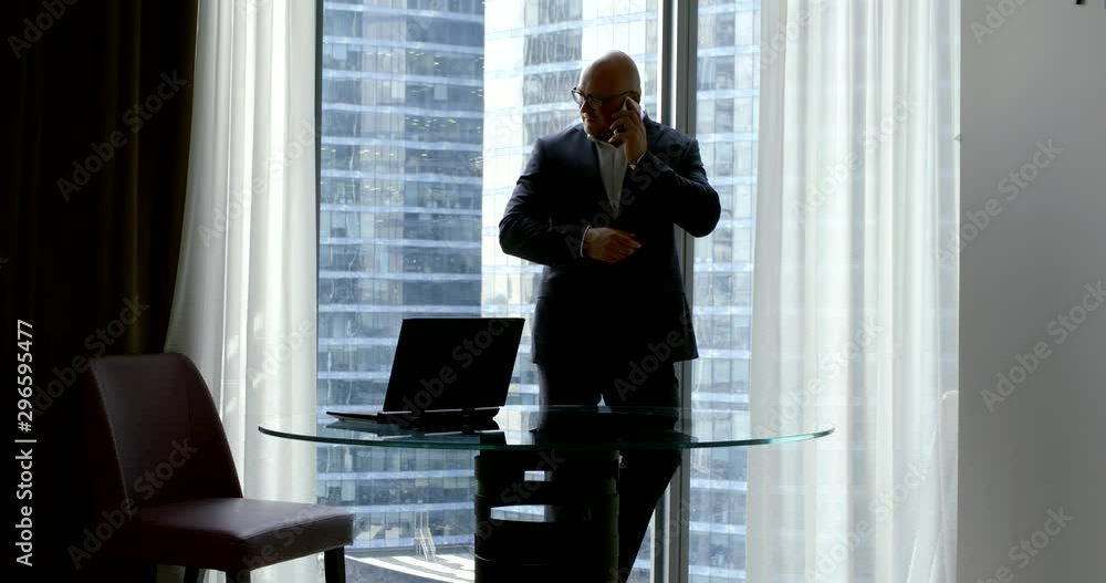 A businessman in a business suit and glasses, he stands in the office at the window in which the skyscrapers are visible near the round glass table on which there is a laptop, talking on the phone. A businessman in a business suit and glasses, he stands in the office at the window in which the skyscrapers are visible near the round glass table on which there is a laptop, talking on the phone.