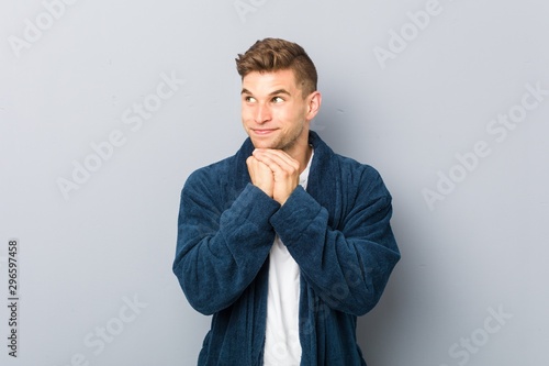 Young caucasian man wearing pajama keeps hands under chin, is looking happily aside.